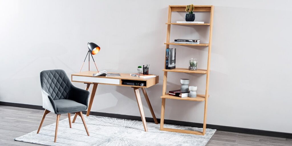 Escritorio de madera clara con silla gris capitoneada y una repisa de escalera con libros y objetos