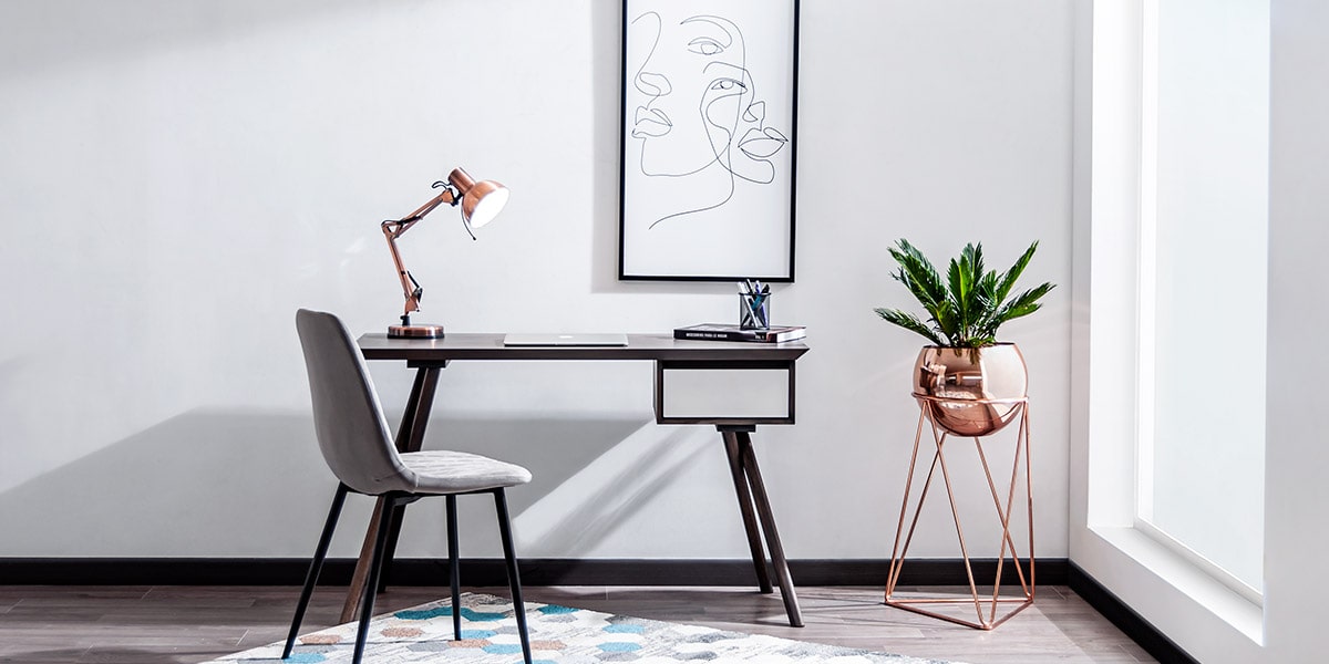 Escritorio oscuro con silla gris, lámpara de cobre y una planta en soporte de cobre, bajo un cuadro de rostro en línea