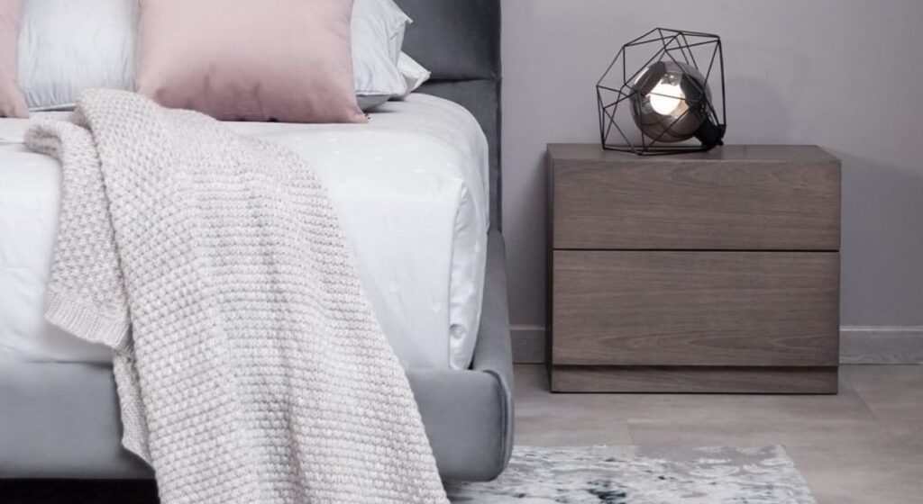 Detalle de una cama con manta tejida clara y cojín rosa junto a una mesa de noche de madera oscura con una lámpara de estructura geométrica