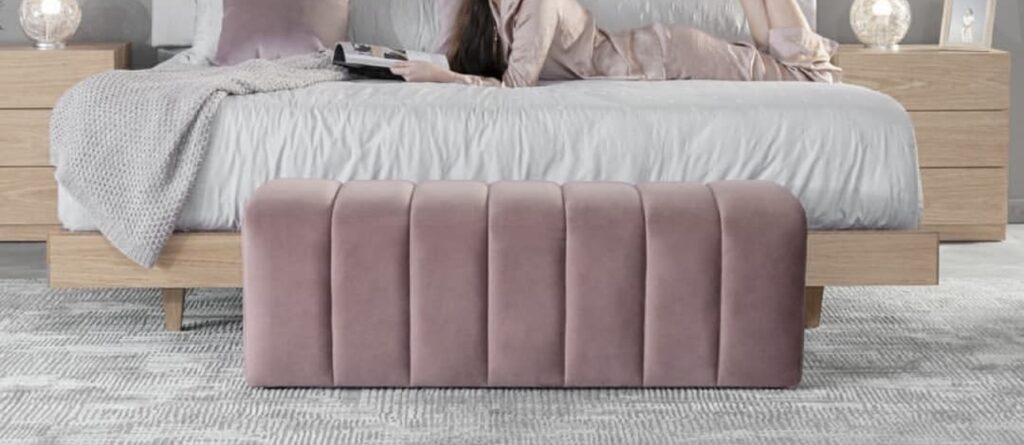 Mujer leyendo una revista sobre una cama con manta gris, con un banco tapizado en rosa palo de canales al pie