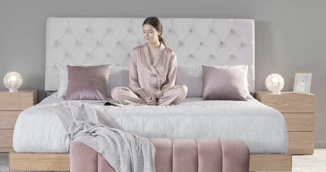 Mujer sentada con las piernas cruzadas sobre una cama con cabecero capitonado blanco, cojines rosa palo, manta gris y un banco rosa al pie