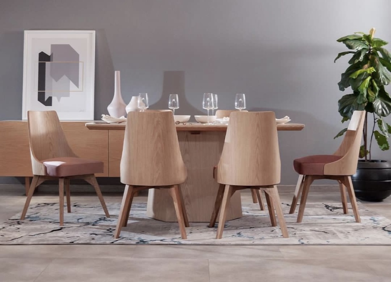 Comedor con mesa de base cónica de madera clara y sillas con respaldo de madera, junto a un aparador, una planta grande y un cuadro abstracto