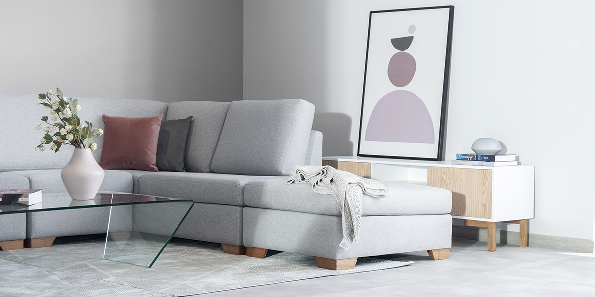 Sofá modular gris claro con chaise longue, mesa de centro de vidrio y un cuadro abstracto, junto a un mueble de madera y blanco