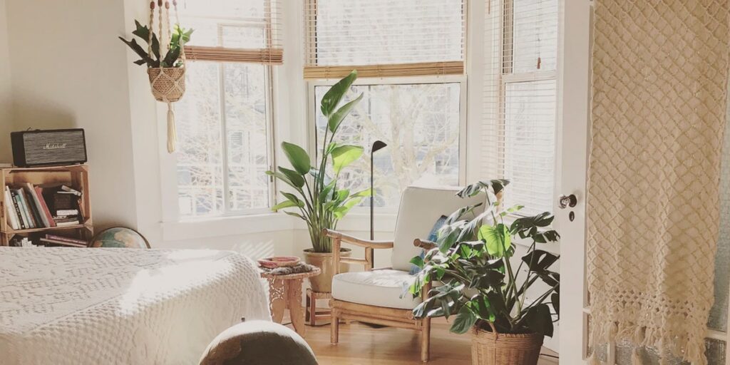 Rincón luminoso con una poltrona de madera, plantas de hojas grandes y una cama con ropa blanca junto a una ventana