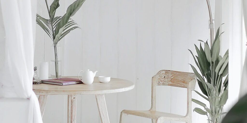 Mesa redonda de madera clara con una tetera, una taza y un libro, junto a una silla de madera y hojas verdes