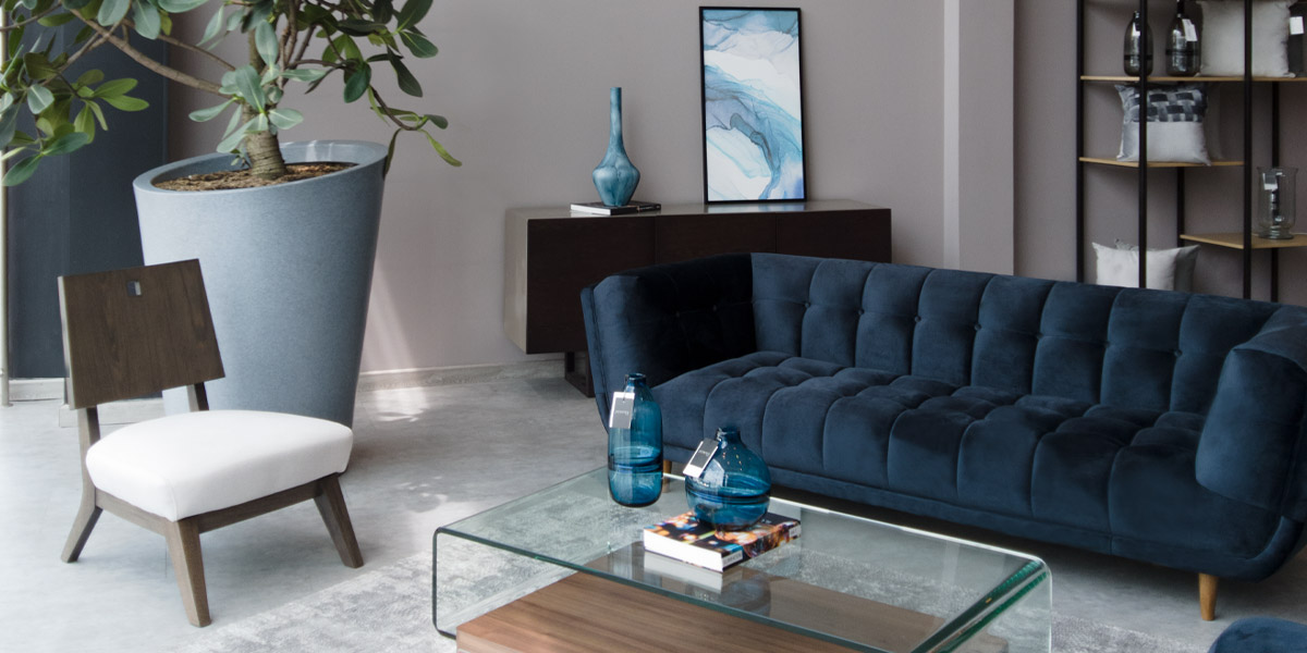 Showroom de Aristas con sofá azul oscuro capitoné, una silla con asiento blanco, mesa de centro de vidrio y una planta en matera