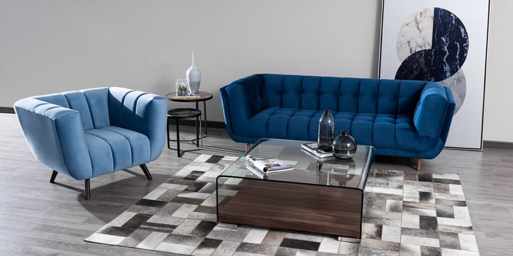 Sofá azul oscuro capitoneado y butaca azul clara de canales con mesa de centro de vidrio sobre un tapete de patchwork en tonos grises