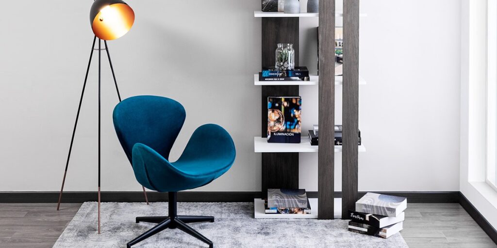 Butaca azul petróleo de líneas curvas bajo una lámpara de trípode, junto a una estantería oscura y blanca con libros