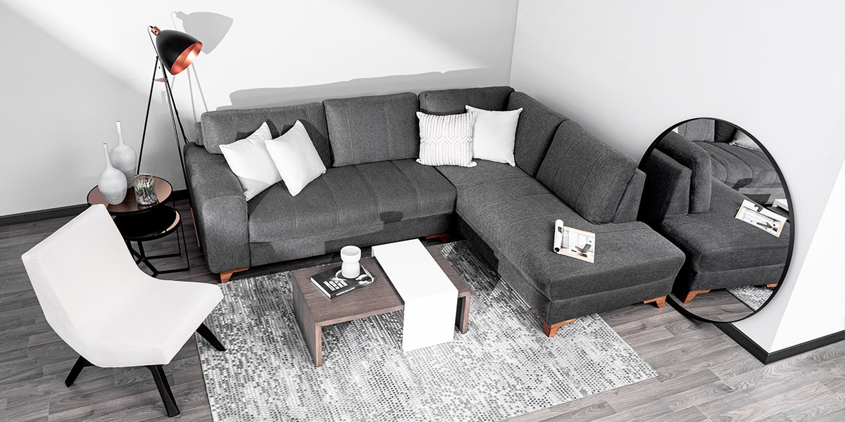 Sofá modular en L gris oscuro con chaise longue, mesa de centro de madera y blanca, y un sillón blanco, junto a un espejo redondo