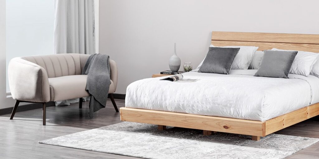 Cama de madera clara con ropa blanca y cojines grises, junto a un sillón crema de líneas curvas con una manta gris