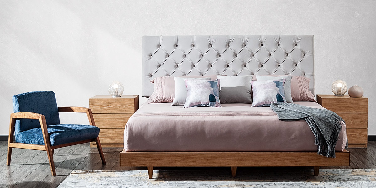 Cama con cabecero capitoné gris claro, ropa rosa y una butaca azul de madera junto a una mesa de noche