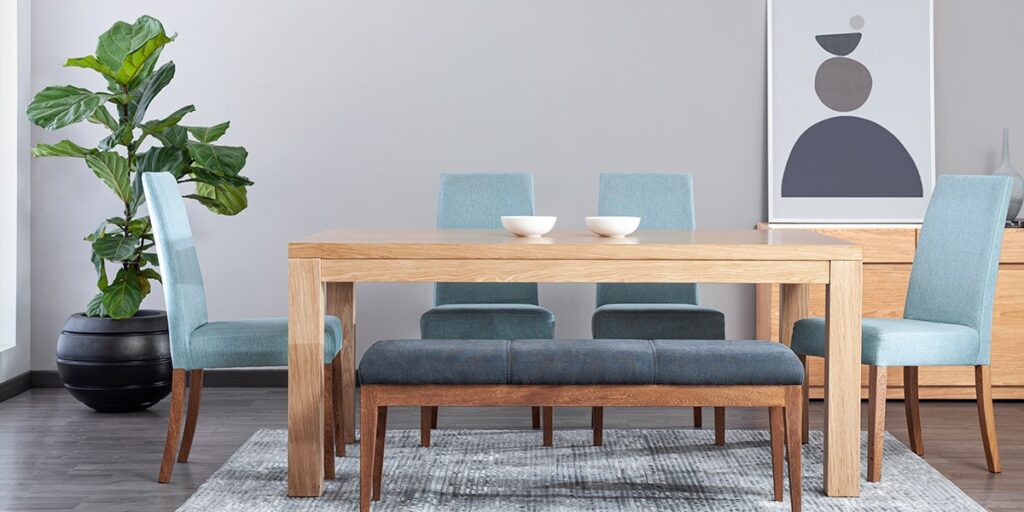 Comedor con mesa de madera clara, sillas azul claro y banca gris azulada, junto a una planta grande en matera negra