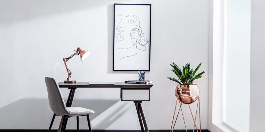 Escritorio de madera oscura con una silla gris, una lámpara de cobre y una planta en soporte de cobre, bajo un cuadro de rostro en línea