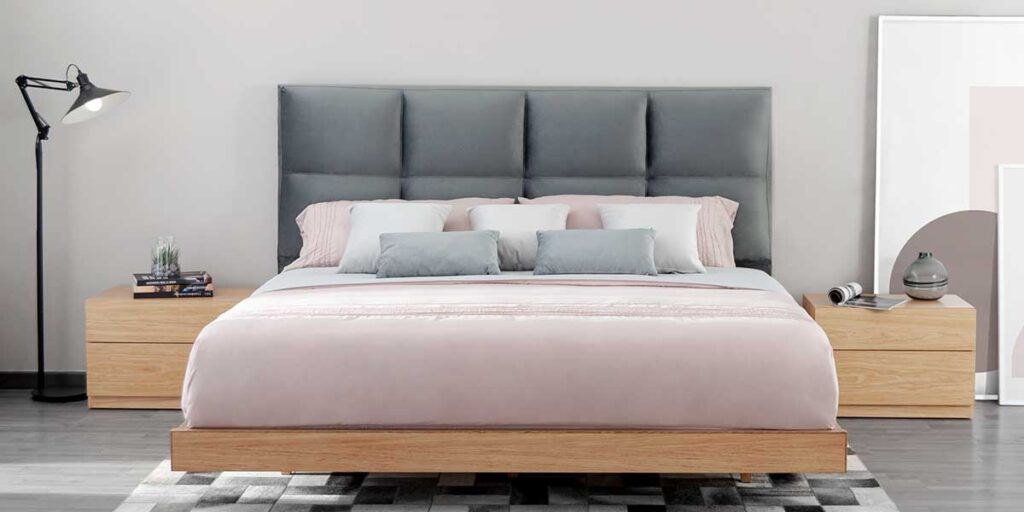 Cama con cabecero gris de paneles, ropa rosa clara y base de madera, con mesas de noche de madera y una lámpara de pie