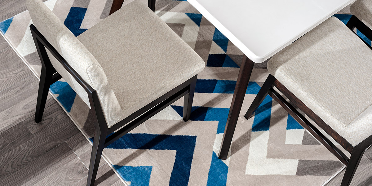 Detalle de dos sillas tapizadas en crema junto a una mesa blanca, sobre un tapete de zigzag azul y gris