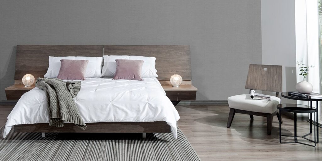 Cama con cabecero y base de madera oscura, ropa blanca, cojines rosa y una manta tejida, con una butaca blanca y mesas de noche