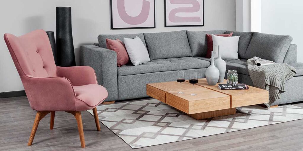 Sala con sofá en L gris, butaca de terciopelo rosa, mesa de centro de madera y tapete geométrico