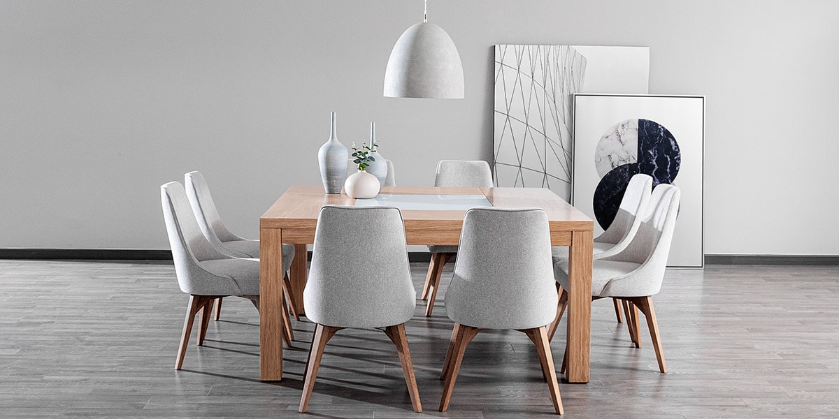 Comedor con mesa de madera clara y sillas grises claras bajo una lámpara colgante blanca, frente a cuadros