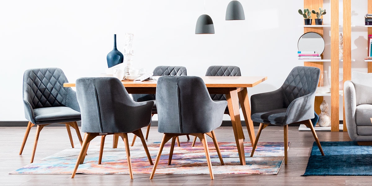 Comedor con mesa de madera y seis sillas tapizadas en gris azulado con capitoné de rombos, sobre un tapete de colores