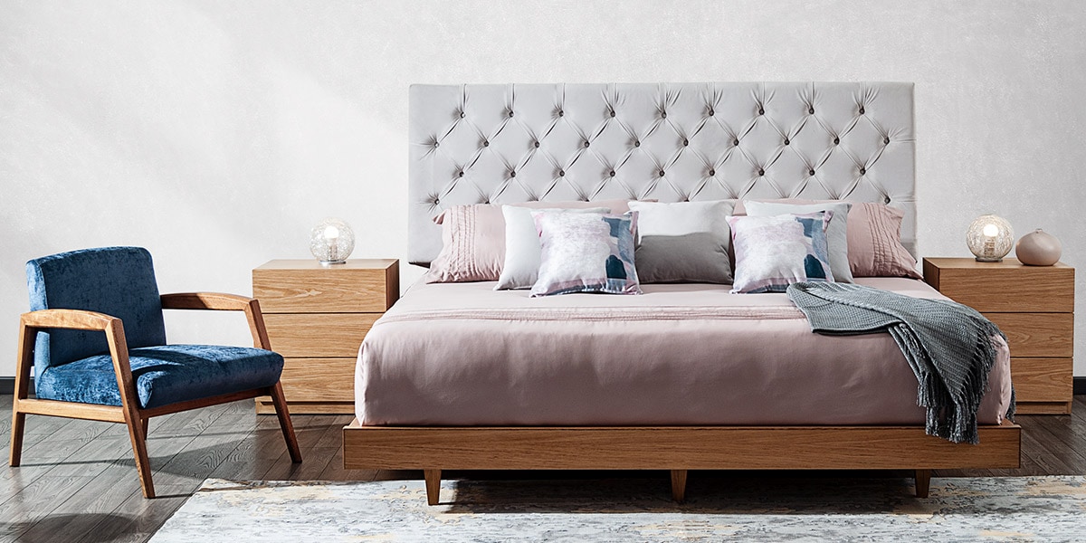 Cama con cabecero capitoné gris claro y base de madera, ropa rosa, una butaca azul y mesas de noche de madera