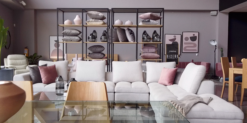 Showroom con sofá modular blanco en L, cojines rosa y gris, mesa de centro de vidrio y estantería metálica con accesorios al fondo