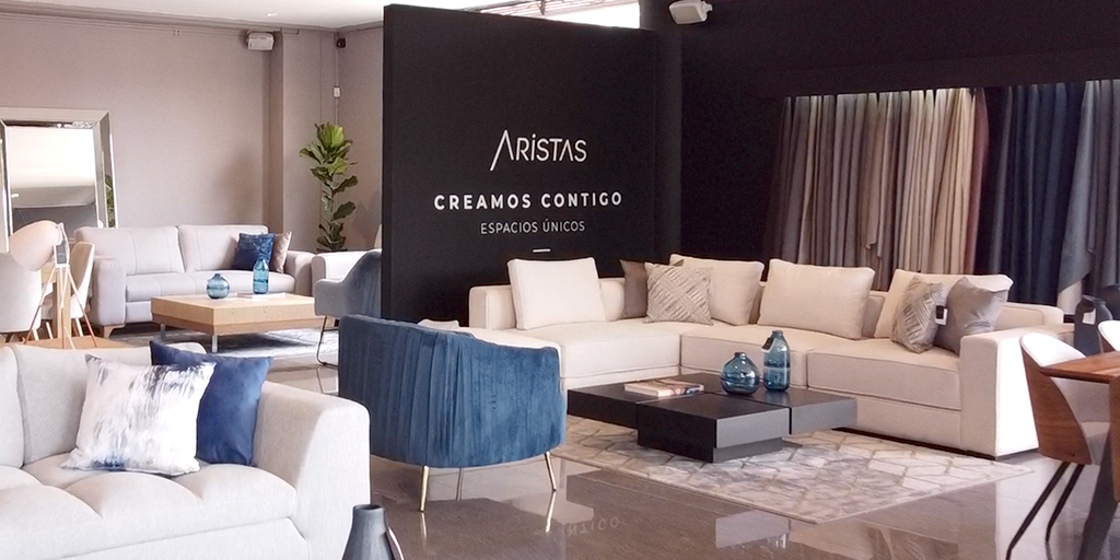 Showroom de Aristas con sala en L en tela blanca, poltrona azul de canales y mesa de centro oscura, frente a un muro negro con el logo «Aristas, creamos contigo espacios únicos»