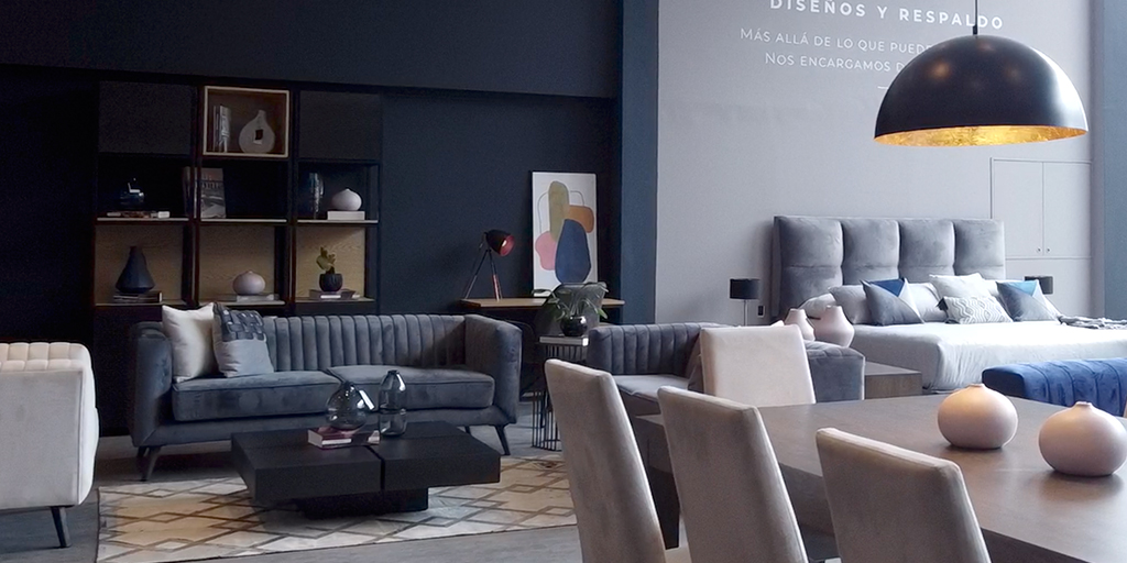 Showroom de Aristas con sofá azul oscuro de canales verticales, mesa de comedor y sillas grises, y una cama con cabecero capitonado gris al fondo