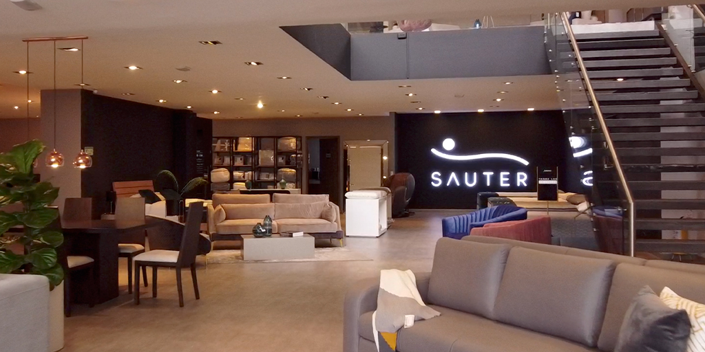Interior de un showroom de dos niveles con escalera, comedores, salas y el logo de Sauter al fondo