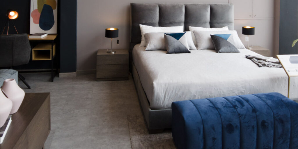 Cama con cabecero gris de paneles, ropa blanca y un banco azul de canales al pie, en un showroom con mesas de noche y lámparas